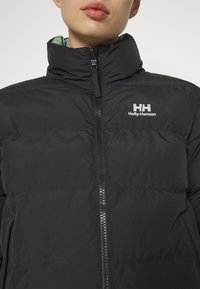 Svart puffjacka med hög krage, framdragkedja och broderad Helly Hansen-logotyp på vänster sida. Slät, quiltad yta.