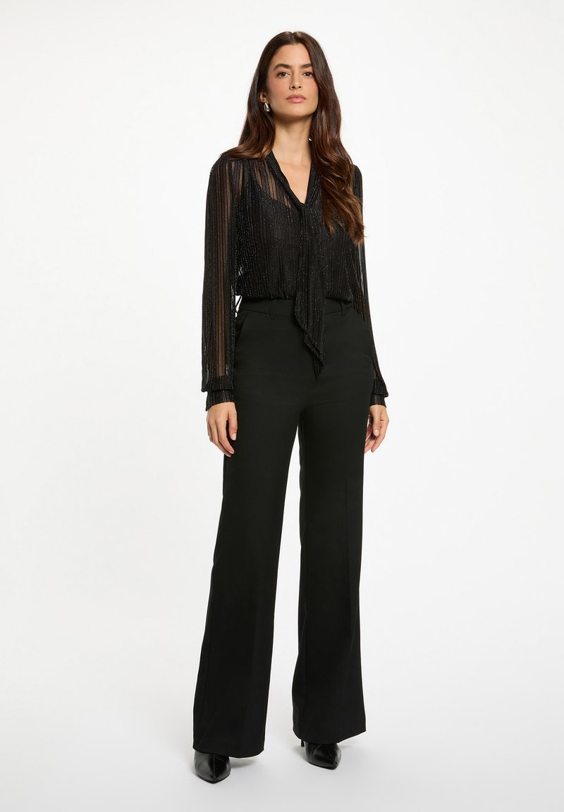 Donna in piedi con una blusa nera trasparente con collo a cravatta e pantaloni neri a vita alta e a gamba larga su uno sfondo bianco.