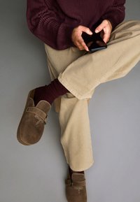 Rjave semiš klompe s spono, v kombinaciji z burgundskimi rebrastimi nogavicami. Kremne hlače in burgundska pulover. Pametni telefon v rokah.