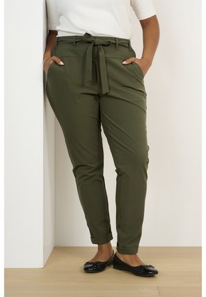 Frau trägt olivgrüne, hoch taillierte Paperbag-Hose mit Gürtel, weißes Oberteil und schwarze Ballerinas, steht mit den Händen in den Taschen.