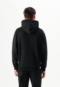 Man wearing plain black hoodie and black pants, standing facing away against a white background.