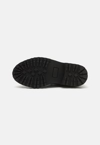 Semelle en caoutchouc noir avec surface texturée, crampons pour traction et une étiquette au niveau du milieu. Conçue pour la durabilité et l'adhérence.