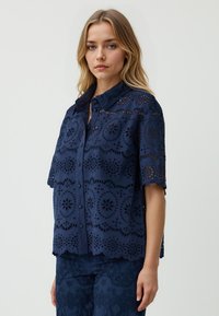 Un modello sta in profilo indossando una blusa ricamata blu navy con maniche corte, caratterizzata da motivi floreali a taglio, su uno sfondo neutro.