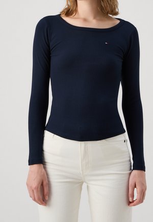 Femme portant un haut bleu marine à manches longues côtelé et ajusté avec un petit logo sur la poitrine, associé à un pantalon blanc taille haute.
