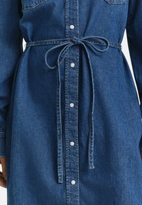 Denim shirt dress, dark blue, featuring button front, long sleeves, waist tie, and front patch pockets with visible stitching detail.