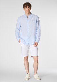 Un giovane uomo si erge con sicurezza, indossando una camicia azzurro chiaro, pantaloni corti bianchi e sneakers, su uno sfondo grigio semplice.