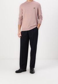 Lichtroze gebreide trui met een ronde hals en een klein geel logo, gecombineerd met zwarte op maat gemaakte broek en zwarte leren schoenen.