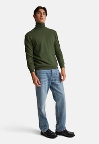 Jersey de cuello alto verde en un material suave, combinado con jeans rectos de color azul claro y zapatos negros, con un acabado suave.