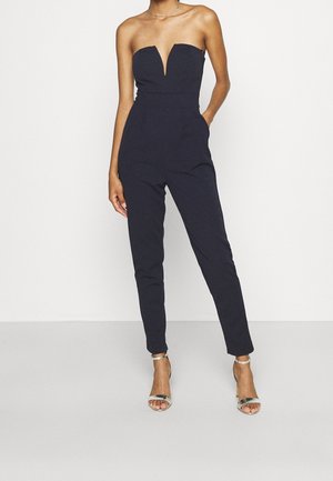 Combinaison bleu marine sans bretelles avec un col en V, des jambes fuselées et des poches latérales. Portée avec des sandales à talons hauts argentées. Texture du tissu lisse.