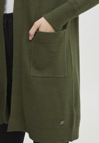 Cardigan vert olive avec des poignets côtelés et des poches. Texture douce et accent logo métallique rond en bas. Design long et ouvert.