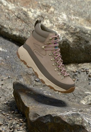 Beige en bruine wandelschoen met roze veters zwevend boven rotsachtig buitengebied, met de nadruk op het robuuste ontwerp en de stevige zool.