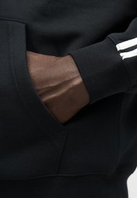 Main à la peau foncée partiellement dans la poche avant d'un sweat-shirt noir avec des bandes blanches sur le bord de la manche.