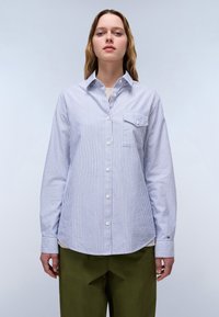 Blue and white striped button-up shirt with a chest pocket, long sleeves, rounded hem, and contrasting cuff patterns. Paired with olive green pants.