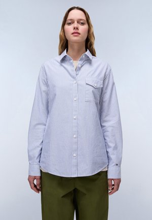 Blue and white striped button-up shirt with a chest pocket, long sleeves, rounded hem, and contrasting cuff patterns. Paired with olive green pants.