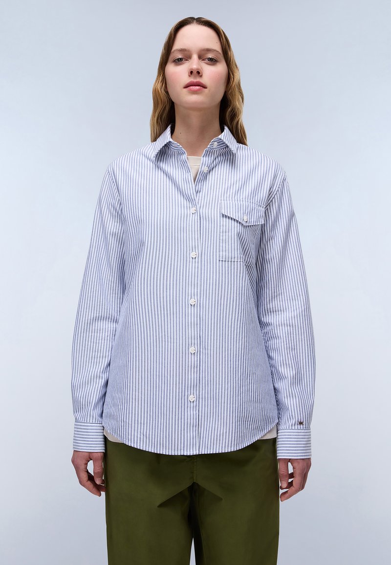 Blue and white striped button-up shirt with a chest pocket, long sleeves, rounded hem, and contrasting cuff patterns. Paired with olive green pants.