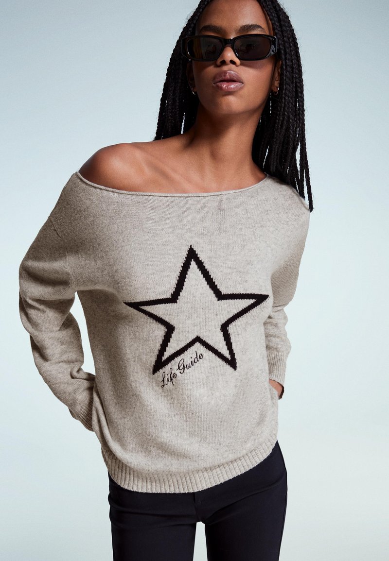 Frau mit geflochtenem Haar, die schwarze Sonnenbrille und einen beigen, schulterfreien Pullover mit einem großen schwarzen Stern und dem Text "Life Guide" trägt.