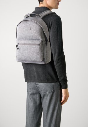Sac à dos en tissu gris avec un dessus arrondi, poche zippée à l'avant, bretelles ajustables et une texture lisse. Porté par une personne en pull sombre.