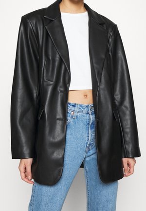 Persona con blazer de cuero negro de gran tamaño sobre top blanco corto y vaqueros azules de tiro alto, mostrada desde el cuello hasta las caderas.