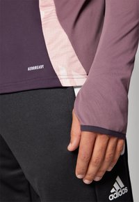 Langarm-Sportshirt in dunklem Lila mit hellrosa Akzenten und strukturiertem Muster, mit einem Logo und Daumenloch an den Ärmeln.