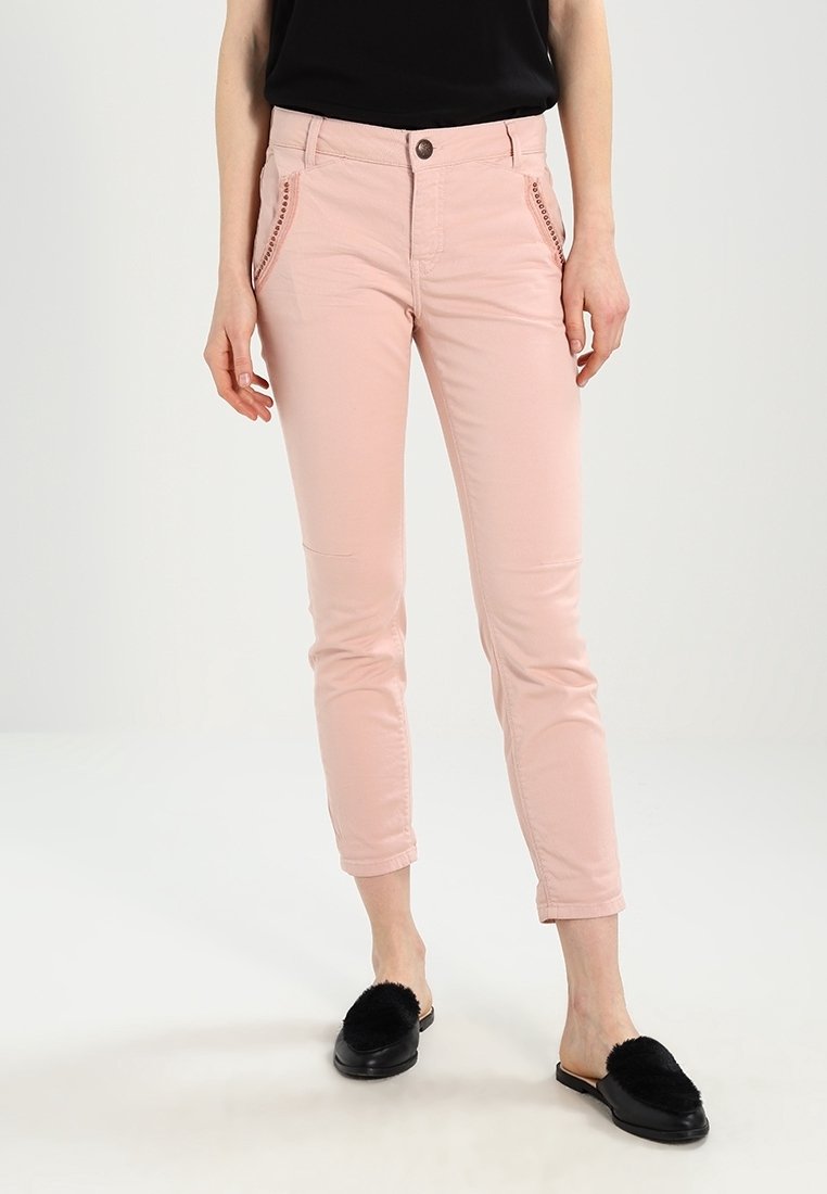 Femme portant un pantalon slim rose clair avec des poches avant cloutées, un haut sans manches noir, et des chaussures noires sans lacets avec un dessus pelucheux, debout.