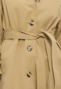 Καμηλό παλτό-τρεντσκότ με ζώνη στη μέση, που διαθέτει τέσσερα μεγάλα κουμπιά και υφή με ανάγλυφη επιφάνεια. Περιλαμβάνει πλαϊνές τσέπες και ραμμένη σιλουέτα.