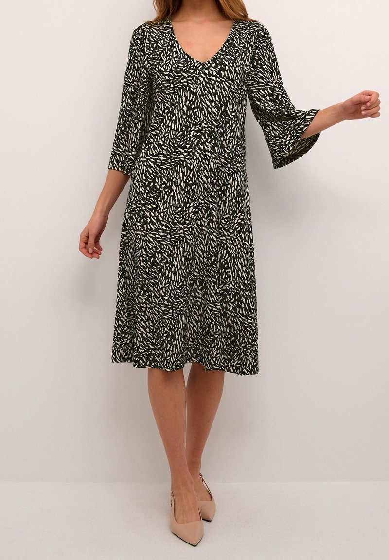 Femme portant une robe aux motifs noir et blanc, longueur genou, manches cloche trois-quarts, et des talons pointus couleur nude.