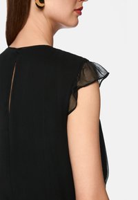 Schwarzes Kleid mit transparenten, flatternden Ärmeln und einem Schlüsselrochen-Rücken. Der Stoff hat eine weiche, strukturierte Oberfläche.