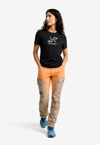 Svart t-shirt med silverlogotyp, orange och beige cargobyxor med sidofickor, och blå atletiska skor. Lockigt hår, gångpose.