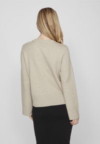 Beige Pullover mit langen Ärmeln und rundem Ausschnitt, mit geripptem Saum. Über einem schwarzen Rock getragen, glatte Textur.