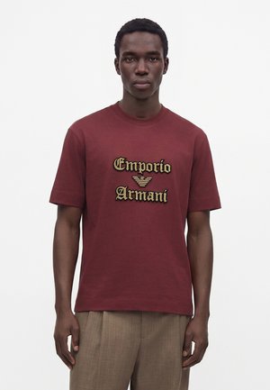 Νέος άνδρας που φοράει μπορντό T-shirt Emporio Armani και μπεζ παντελόνια, στέκεται μπροστά σε μια λευκή απλή φόντο.