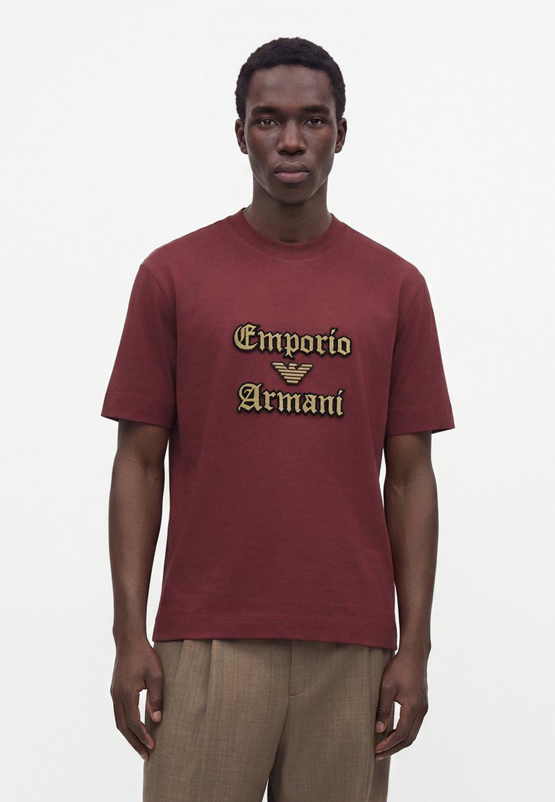 Νέος άνδρας που φοράει μπορντό T-shirt Emporio Armani και μπεζ παντελόνια, στέκεται μπροστά σε μια λευκή απλή φόντο.