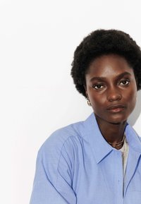 Chemise bleu clair à boutons avec un tissu texturé, dotée d'un col traditionnel et de manches courtes, portée au-dessus d'un haut gris.