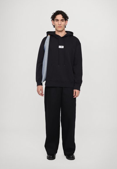 Sweatshirt à capuche noir avec poche avant et étiquette graphique, associé à un pantalon noir ample et des chaussures noires. Accent en denim sur l'épaule.