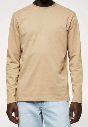 Dunkelhäutige Person, die ein schlichtes beigefarbenes Langarmshirt und hellblaue Jeans trägt, vor einem einfarbigen Hintergrund steht.
