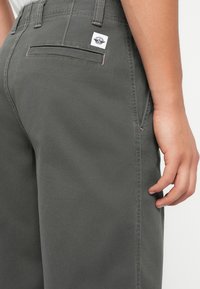 Dark grey cotton pants with a rear pocket and a white brand label. Contrast stitching in lighter pink is visible along the seams.