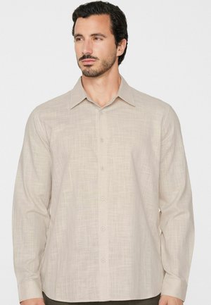 Mann mit dunklem Haar und Bart, der ein beiges langärmliges Hemd mit Knöpfen trägt und leicht nach links blickt vor schlichtem Hintergrund.