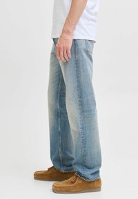 Hombre vestido con jeans rectos de color azul claro, una camiseta blanca y zapatos informales de gamuza marrón, de pie contra un fondo liso.