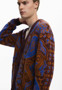 Cardigan marrón con patrones azules en espiral, presenta un escote en V profundo, botones frontales y puños acanalados. Material de punto suave con textura.