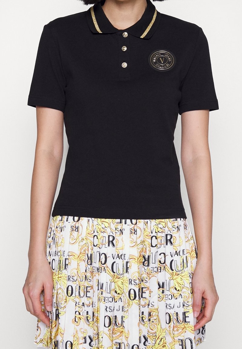 Vrouw draagt een zwart poloshirt met gouden bies en knoopdetails, gecombineerd met een witte geplooide rok met een patroon van zwarte tekst en gele ontwerpen.