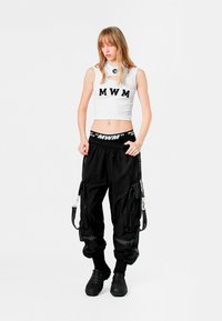 Top corto blanco con la letra "MWM", combinado con pantalones cargo holgados negros que tienen cremalleras y correas, y calzado negro texturizado.