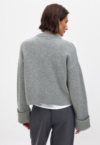 Apsēts pelēks adīts džemperis ar ribotu apkakli un aprocēm, maiga tekstūra, brīva piegriezuma, kopā ar pelēkiem biksēm.