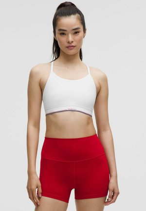 Weißer Sport-BH mit dünnen Trägern, versehen mit einem rot-blauen gestreiften Bund. Kombiniert mit hochgeschnittenen roten Shorts, beide aus glattem Material gefertigt.