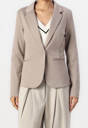 Femme portant un blazer taupe ajusté sur un haut blanc à encolure en V festonnée et un pantalon plissé beige taille haute.