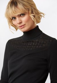 Schwarzes Langarmoberteil mit hohem Kragen, das ein transparentes, dekoratives Spitzendesign im oberen Brustbereich und ein weiches Strickdesign aufweist.
