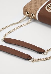 Angolo di una borsa a tracolla marrone con motivi, dotata di catene dorate e spalline in pelle, posata su una superficie bianca.