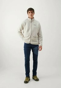 Chaqueta de forro polar blanca con cremallera y cuello alzado, dos bolsillos frontales, llevada sobre pantalones azul marino y zapatos verde oliva. Tela simple y texturizada.