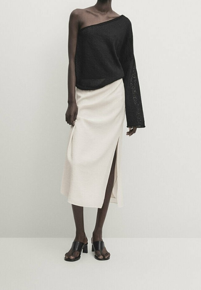 Personne à la peau foncée portant un top noir asymétrique à manches longues, une jupe midi blanche avec une fente latérale et des sandales à talons noires ouvertes à l'avant.