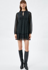 Schwarzes, durchsichtiges Kleid mit weißen Punkten, gerafftem Oberteil, Schlüssellochausschnitt und langen Ärmeln. Kombiniert mit knielangen schwarzen Lederstiefeln.