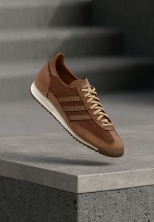 Brun ruskind- og tekstilsneaker med vævet struktur, med lyse beige snørebånd og tre karakteristiske striber på siden. Gummisål.