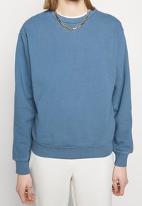 Sweat-shirt bleu à col rond avec manches longues et poignets et ourlet côtelés, fabriqué en matériau doux. Porté avec une chaîne en or.